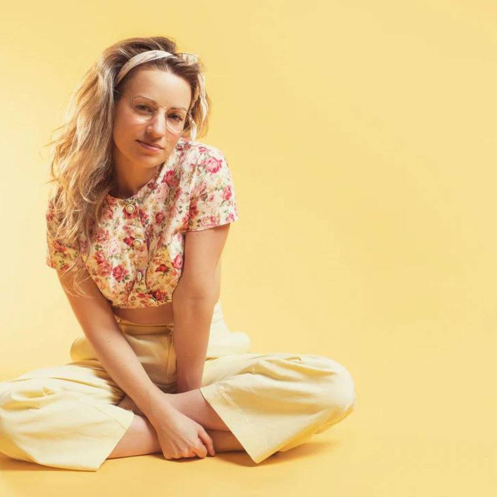 Women business owner in yellow pants, yellow/red floral shirt sitting crosslegged. White background.